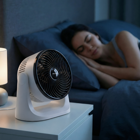 Woman sleeping in bed with a fan on a nightstand next to her.