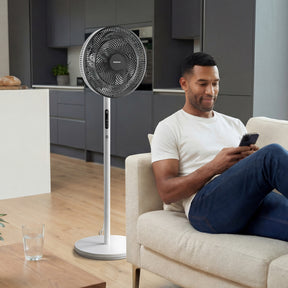 Man sitting on a couch using a smartphone with a standing fan in the foreground.