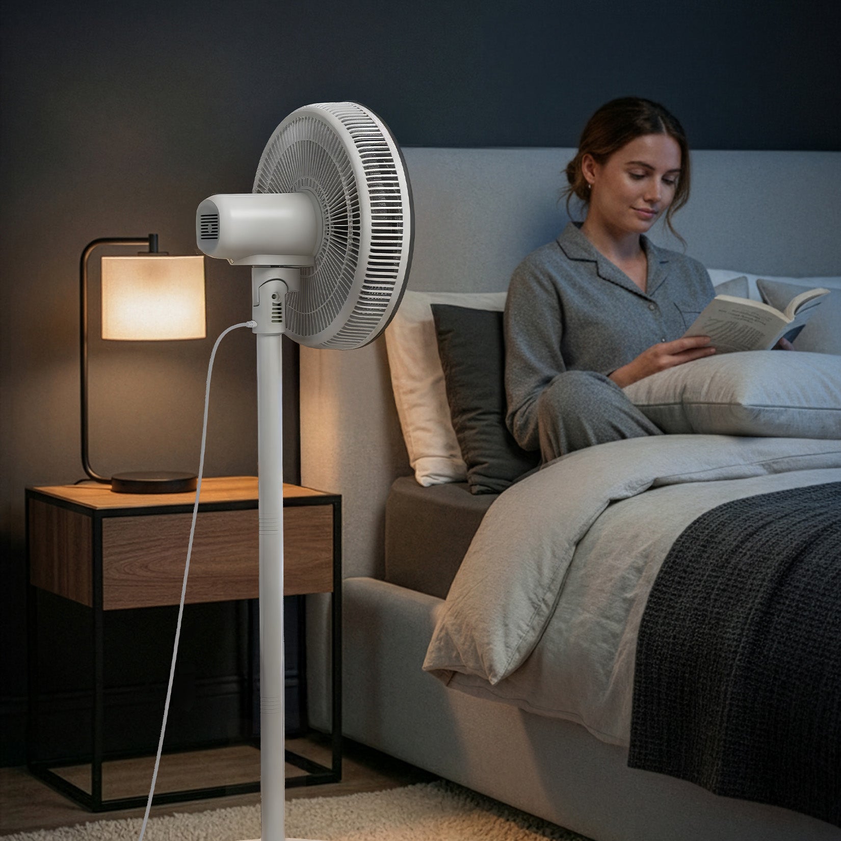 Woman reading a book in bed next to a standing fan and lamp.